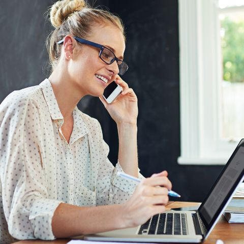 Photo of woman with glasses working on laptop while on the phone.